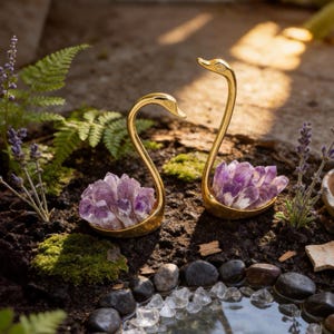 May include: Two gold-colored swan figurines, each cradling a cluster of purple amethyst crystals. The swans are set in a garden setting with plants and a small pond, creating a decorative and natural aesthetic.