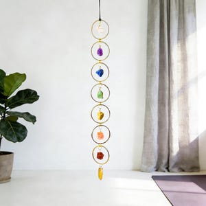 Op de afbeelding: Een verticale hangende decoratie met zeven goudkleurige metalen ringen in een kolom. Elke ring bevat een enkele ruwe, ongeslepen edelsteen in verschillende kleuren, waaronder wit, paars, blauw, groen, geel, oranje en rood.