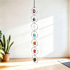 May include: A vertical hanging decoration featuring seven silver-toned metal rings linked together in a chain. Each ring contains a single raw, wire-wrapped gemstone in various colors including purple, black, clear, green, orange, red, and dark gray.