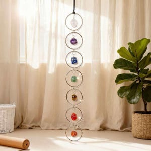 Op de afbeelding: Een hangende chakra kristallen mobiel met zeven verschillend gekleurde ruwe edelstenen, opgehangen in zilveren metalen ringen. De edelstenen zijn helder, paars, blauw, groen, bruin, rood en oranje. De mobiel hangt aan een zwart koord.