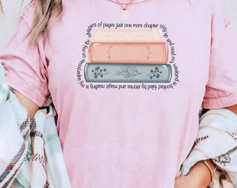 Puede incluir: Camiseta rosa pálido con un gráfico de tres libros apilados en tonos pastel. El texto alrededor de los libros dice: "mi superpoder es leer, alimentado por historias y magia, relájate y lee mi fin de semana".