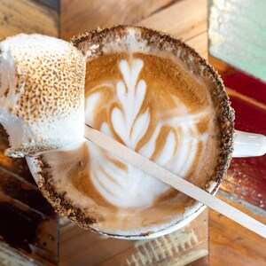 Może przedstawiać: Biały ceramiczny kubek z brązową obwódką wypełniony latte z wzorem liścia. Pieczone pianki są częściowo zanurzone w latte.