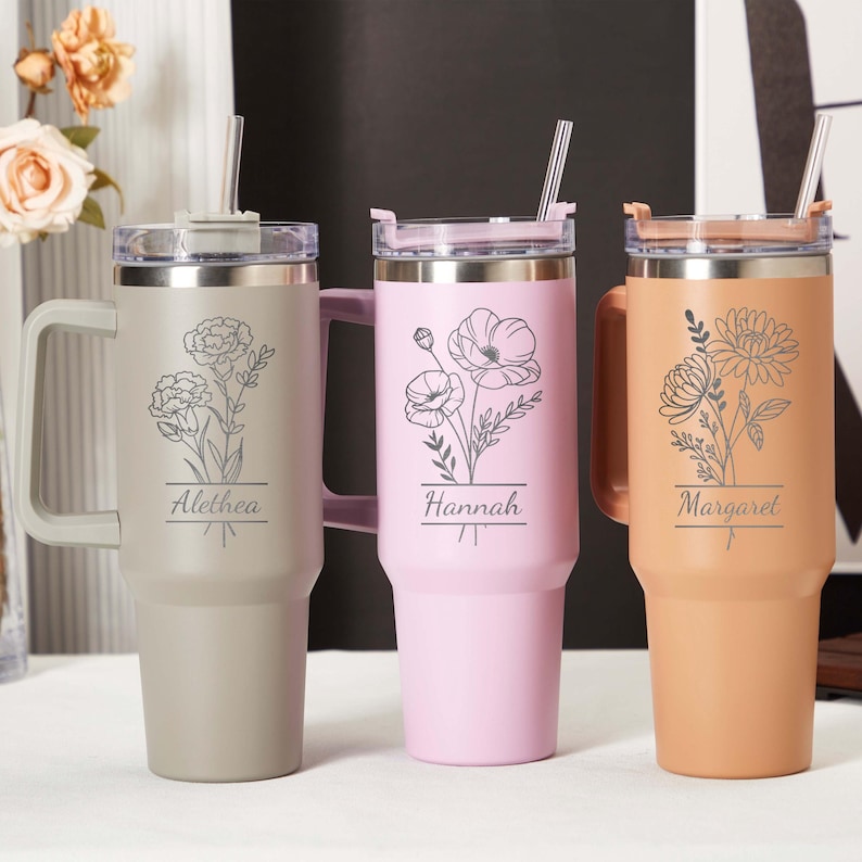 May include: Three insulated tumblers in beige, pink, and peach, each with a handle, straw, and floral engraving. The beige tumbler has the name "Alethea", the pink one "Hannah", and the peach one "Margaret".