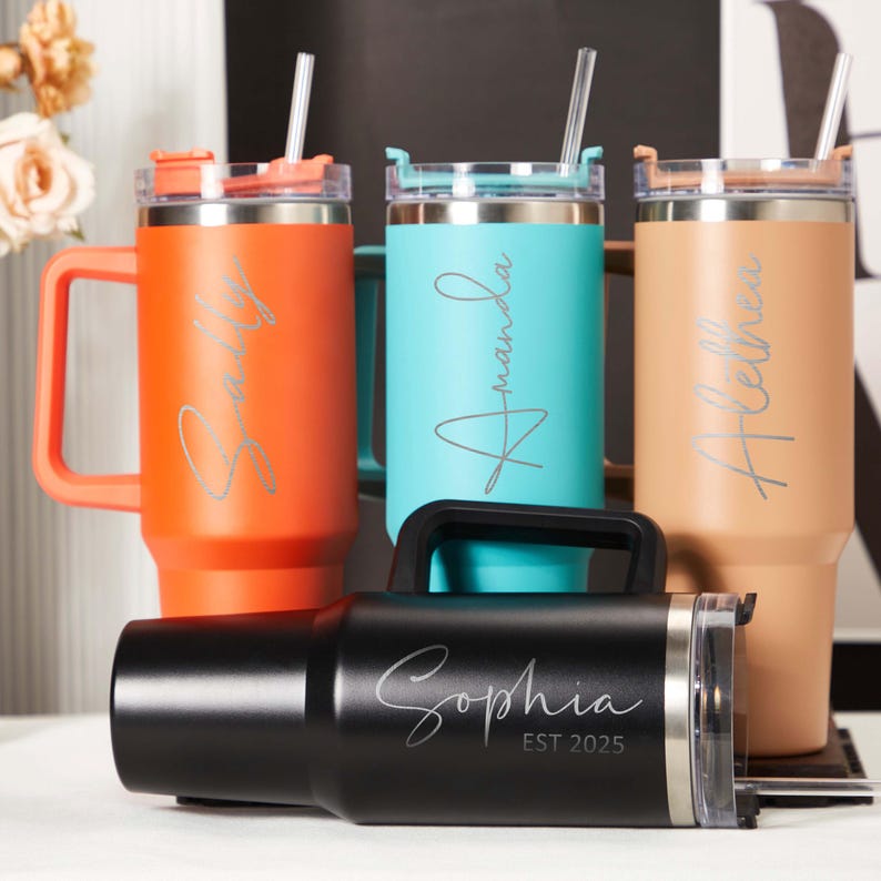 May include: An assortment of personalized tumblers in various colors, including orange, teal, and tan. Each tumbler features a name in a cursive font. One black tumbler has the name "Sophia" and the text "EST 2025". All tumblers have handles and straws.