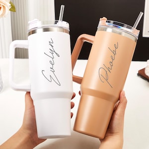May include: Two personalized tumblers with handles. One is white with "Evelyn" in script, the other is tan with "Phoebe" in script. Both have clear lids, stainless steel rims, and clear straws. The tumblers are designed to keep drinks cold.