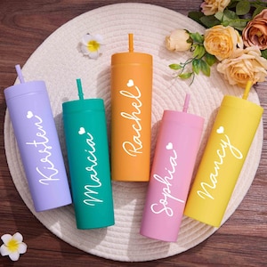 Puede incluir: Cinco vasos de colores con pajitas, cada uno de un color diferente: morado, verde azulado, naranja, rosa y amarillo. Cada vaso tiene un nombre en escritura blanca y un pequeño corazón. Los vasos están colocados sobre una esterilla blanca tejida.