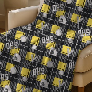 May include: A black, gray, and yellow plaid throw blanket draped over a beige armchair. The blanket features a repeating pattern of baseball helmets, bats, balls, and the text "OHS" in white.
