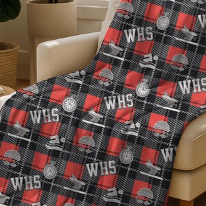 May include: A red, black, and gray plaid blanket draped over a beige armchair. The blanket features repeating patterns of a stopwatch, a hat, and the letters "WHS" in white. The blanket is made of a soft material.