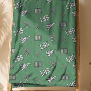 May include: A green blanket with a repeating pattern of the letters "LHS", pennants that say "SCORE", and lacrosse sticks. The blanket is draped over a wooden ladder. The background includes a vase with dried plants and a small wooden stool.