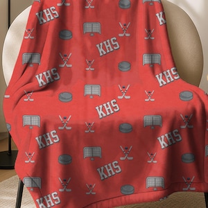 May include: A red hockey-themed blanket draped over a chair. The blanket features a repeating pattern of hockey pucks, nets, and sticks, along with the letters "KHS" in white. The blanket is made of a soft material.