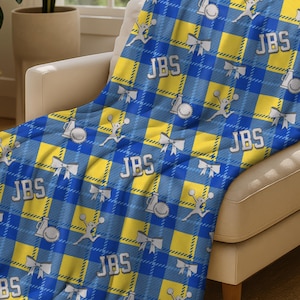May include: A blue and yellow plaid throw blanket draped over a beige armchair. The blanket features a repeating pattern of the initials "JBS", bows, and other decorative elements. The blanket is made of a soft material and is suitable for indoor use.