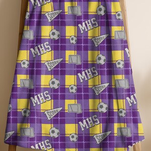 May include: A purple and yellow plaid blanket with a soccer theme. The blanket features soccer balls, goals, and the text "MHS" and "SCORE" in white. The blanket is draped over a wooden ladder.