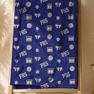 May include: A navy blue blanket with a repeating pattern of volleyballs, bows, and the letters "PHS" in white. The blanket is draped over a wooden ladder, with a neutral background.