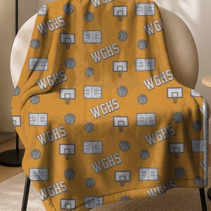 May include: Mustard yellow throw blanket with a repeating pattern of basketballs, basketball hoops, and basketball courts. The text "WGHS" is also repeated throughout the design. The blanket is draped over a chair.