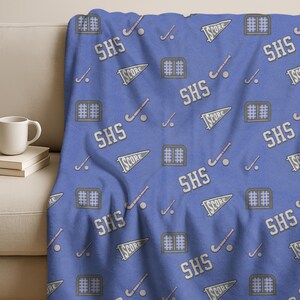 May include: A blue blanket with a repeating pattern of hockey sticks, pucks, pennants that say "SCORE", and the letters "SHS". The blanket is draped over a white couch. The blanket is made of a soft material.