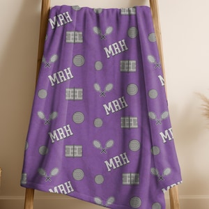 May include: A purple tennis-themed blanket draped over a wooden ladder. The blanket features a pattern of tennis balls, rackets, and court designs, along with the initials "MAH" in white. The blanket is a vibrant and sporty accessory.