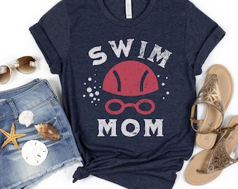 Könnte beinhalten: Marineblaues T-Shirt mit dem weißen Aufdruck "SWIM MOM" und einer roten Schwimmkappe und Schwimmbrillen-Grafik. Das Bild zeigt Jeansshorts, Sonnenbrille und Sandalen, was einen sommerlichen Look erzeugt.
