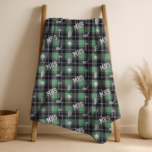 May include: A black, green, and white plaid golf-themed blanket with the initials "MHS" repeated throughout. The blanket features golf balls, clubs, and tees. The blanket is draped over a wooden ladder.