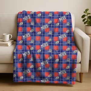 May include: A blue, red, and white plaid throw blanket with footballs, helmets, and the letters "MHS" repeated across the surface. The blanket is draped over a white sofa.