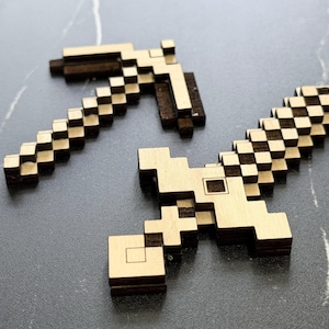 May include: Two wooden pixelated toys: a pickaxe and a sword. The pickaxe has a brown handle and a light brown head. The sword is light brown with a checkered pattern on the blade. The toys are on a dark gray surface.