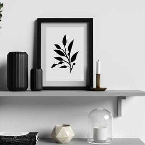 May include: A black framed print with a minimalist black and white botanical design. Two black cylindrical vases, a brass candle holder with a white candle, and a geometric vase are on a gray shelf. A glass cloche with a candle is also present.