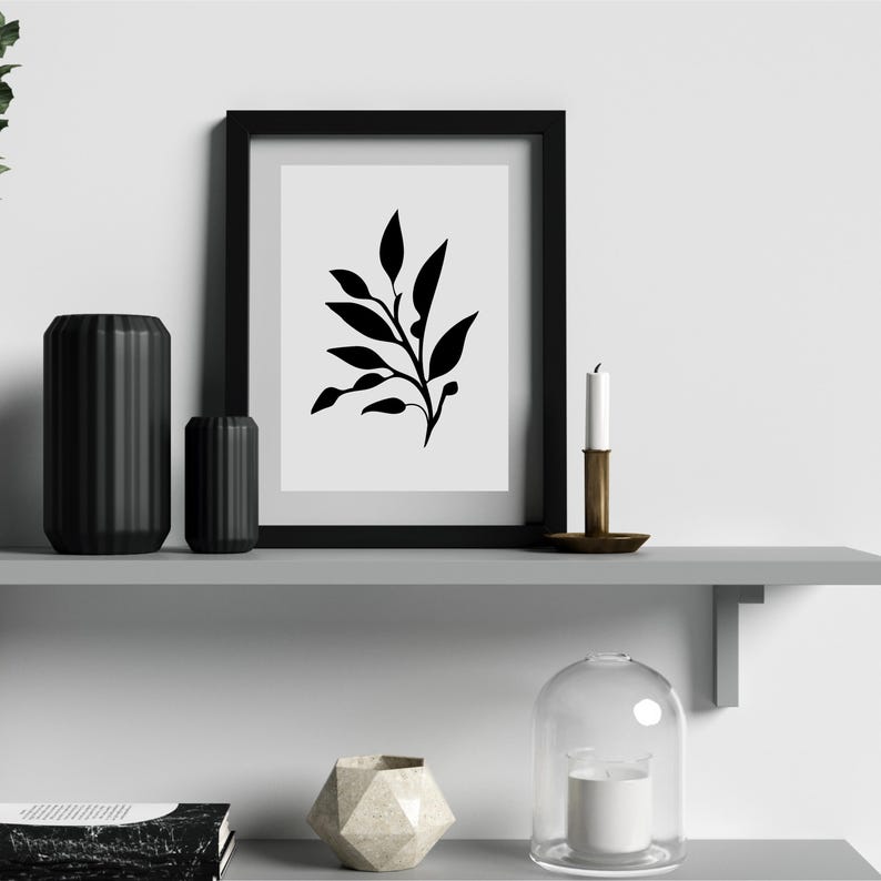 May include: A black framed print with a minimalist black and white botanical design. Two black cylindrical vases, a brass candle holder with a white candle, and a geometric vase are on a gray shelf. A glass cloche with a candle is also present.