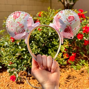 May include: Floral-patterned mouse ear headband with pink satin bows and a sparkling gem. The ears are made of fabric with a patchwork design featuring red roses and green leaves. The headband is held up by a hand.
