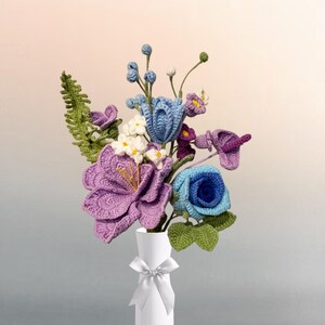 Puede incluir: Un ramo de flores de ganchillo en un jarrón blanco. El ramo incluye flores moradas, azules y blancas con hojas verdes.