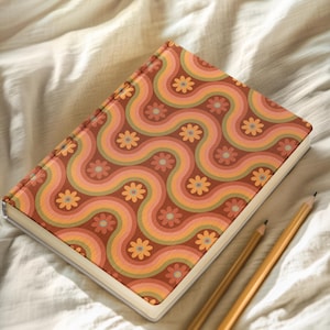 May include: A hardback journal with a retro floral pattern in shades of orange, pink, and green. The cover design features wavy lines and flower motifs. Two pencils rest beside the journal on a white surface.