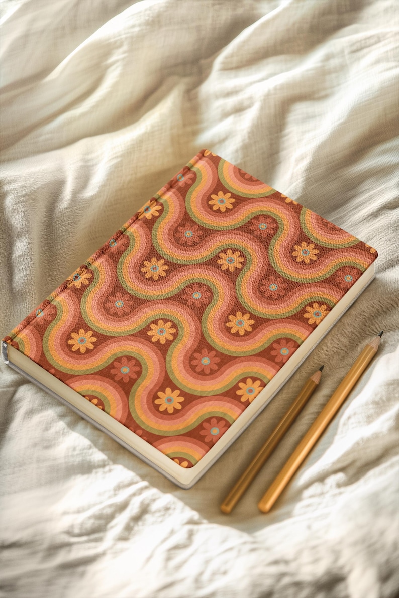 May include: A hardback journal with a retro floral pattern in shades of orange, pink, and green. The cover design features wavy lines and flower motifs. Two pencils rest beside the journal on a white surface.