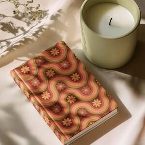 May include: A decorative notebook with a retro floral pattern in shades of brown, orange, and pink. A light green candle sits next to the notebook. The candle has a white wax and a black wick. The items are on a cream-colored surface.