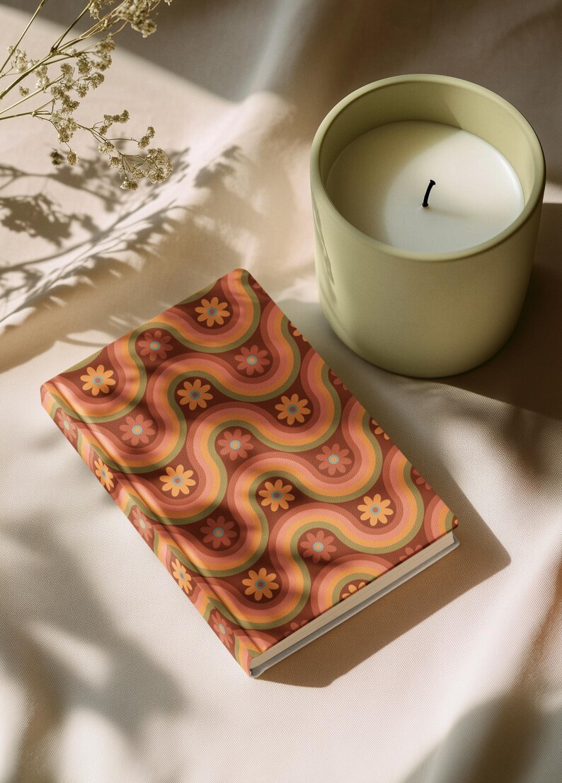 May include: A decorative notebook with a retro floral pattern in shades of brown, orange, and pink. A light green candle sits next to the notebook. The candle has a white wax and a black wick. The items are on a cream-colored surface.