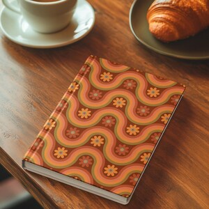 May include: A hardback notebook with a retro-style wavy pattern in shades of brown, orange, and pink, adorned with small flower motifs. The notebook is on a wooden table, next to a cup of coffee and a croissant.