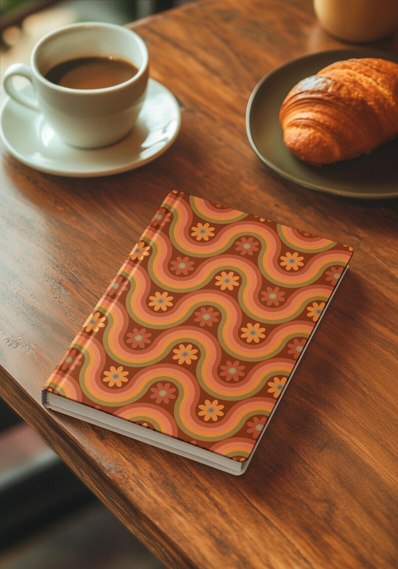 May include: A hardback notebook with a retro-style wavy pattern in shades of brown, orange, and pink, adorned with small flower motifs. The notebook is on a wooden table, next to a cup of coffee and a croissant.