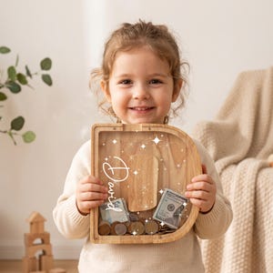 Puede incluir: Una hucha de madera con forma de letra D y panel frontal transparente. El nombre David está impreso en caligrafía blanca sobre el cristal, rodeado de pequeños gráficos de estrellas blancas. El interior contiene monedas y un billete.