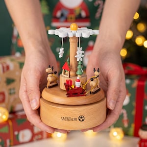 Op de afbeelding: Een houten kerstcarrousel muziekdoos met een centrale zuil, een witte sneeuwvlok als bovenkant en draaiende figuren waaronder rendieren, een rode slee, een kerstman en groene bomen. De voet is gegraveerd met de naam William en een voetbal.