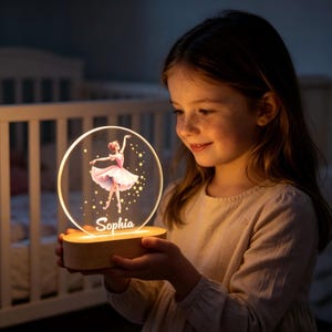 May include: A round, illuminated night light featuring a ballerina in a pink tutu and a name, "Sophia". The light is set on a wooden base. The ballerina is surrounded by gold stars. The night light is held by a person.