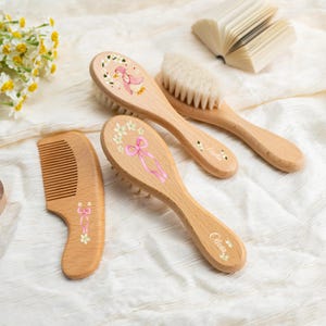 Könnte beinhalten: Ein hölzernes Babypflegeset mit Kamm und Bürsten. Die Bürsten haben weiche Borsten und sind mit bemalten Blumen- und Schleifendesigns verziert. Eine Bürste hat eine rosa Entenillustration, und eine andere hat den Namen "Olivia" darauf stehen.