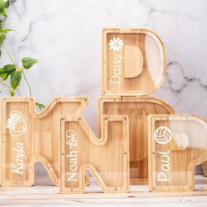 Può includere: Salvadanai in legno a forma di lettera con frontali in acrilico trasparente. Le lettere compongono le parole "KIND" e "P", con nomi ed elementi decorativi come un fiore e un pallone da pallavolo. I salvadanai sono in legno chiaro.
