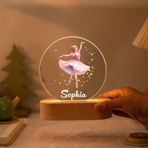 Może przedstawiać: Spersonalizowana lampka nocna z baletnicą w różowej sukience, gwiazdami i imieniem "Sophia". Światło umieszczone jest na drewnianej podstawie. Przedmiot dekoracyjny.