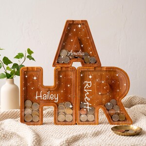 May include: Three wooden letter-shaped coin banks in the forms of A, H, and R. Each letter features a clear front panel, internal metal coins, and white star graphics. The names Amelia, Hailey, and Rhett are printed in white script on the front.