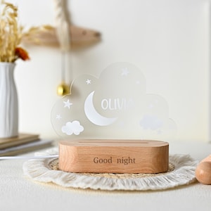 Puede incluir: Luz de noche de madera con un panel acrílico en forma de nube. El panel presenta una luna creciente, estrellas y el nombre "OLIVIA". La base de madera está grabada con "Good night". Para un dormitorio infantil.