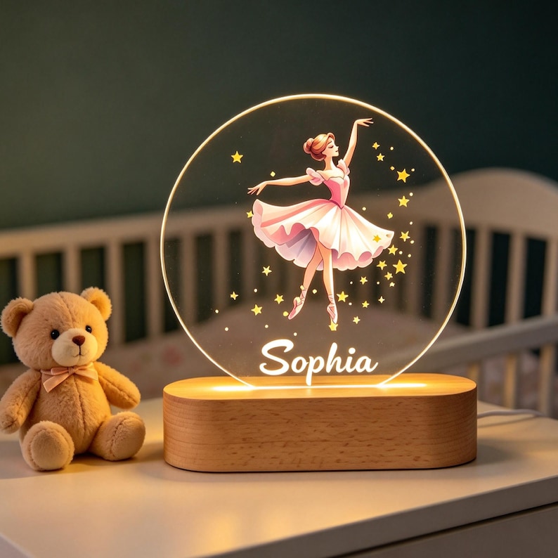 Może przedstawiać: Okrągła lampka nocna z akrylu z ilustracją baletnicy w r&oacute;żowej sukience, otoczonej ż&oacute;łtymi gwiazdkami. Imię Sophia jest nadrukowane białą czcionką pod tancerką. Lampka umieszczona jest na podstawie z jasnego drewna.