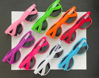 May include: Nine pairs of colorful sunglasses with personalized names printed on the arms. The sunglasses are in various colors, including pink, purple, green, orange, red, white, and blue.
