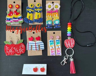 Könnte beinhalten: Eine Auswahl an Lehrer-Themed-Ohrringen aus Perlen, ein Perlenband mit einem roten Apfel-Anhänger, auf dem "Teacher" steht, und ein Schlüsselanhänger mit einem roten Apfel-Anhänger. Die Ohrringe haben Designs wie Äpfel, Bücher, Katzen und die Wörter "Teach" und "Fri-Yay".
