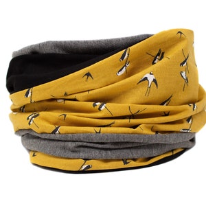 Op de afbeelding: Een gele nekwarmer met een zwart-wit vogelpatroon. De nekwarmer is gemaakt van een zachte, rekbare stof en is omkeerbaar. Hij heeft een grijze laag aan de binnenkant.