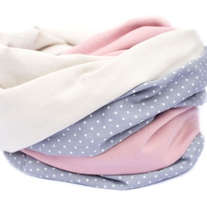 May include: A soft, cozy neck warmer made of three different fabrics: a light pink, a light gray with white polka dots, and a cream color. The neck warmer is folded and rolled up.