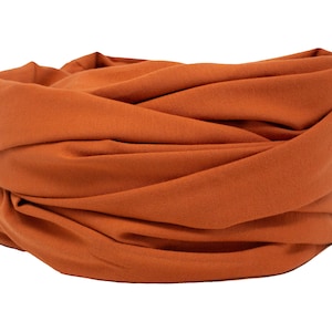 Puede incluir: Un tubo de tela de color naranja quemado, probablemente una diadema o un calentador de cuello, se muestra sobre un fondo blanco. El material es suave y parece estar doblado o envuelto, creando un efecto de capas. El color es un tono cálido y terroso.