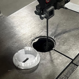 May include: A bandsaw with a silver blade and a gray metal table. A light gray, circular, 3D-printed part sits on the table. The bandsaw is in a workshop setting.