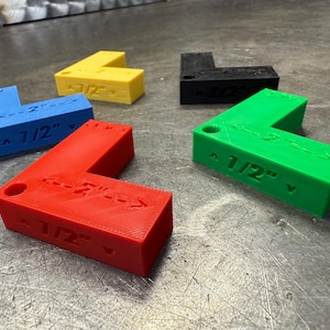 Könnte beinhalten: Fünf L-förmige Eckwerkzeuge in den Farben Rot, Blau, Gelb, Grün und Schwarz. Jedes Werkzeug hat die Maße "1/2" und "2" auf seinen Seiten. Die Werkzeuge bestehen aus einem festen Material und sind für präzise Winkelmessungen konzipiert.